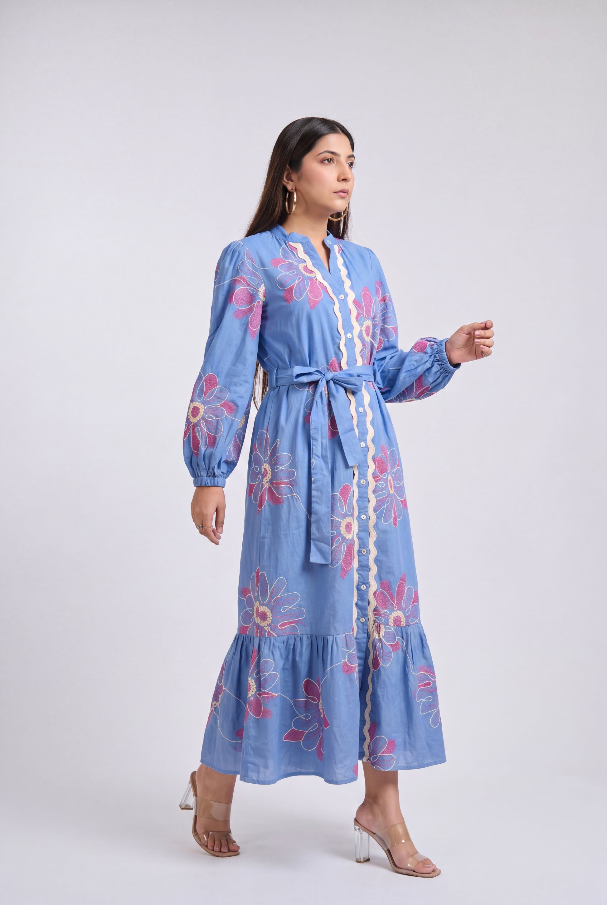 A woman modeling a light blue long-sleeve maxi dress with pink and white floral embroidery and a cream-colored tie detail down the front, set against a plain light gray studio background.