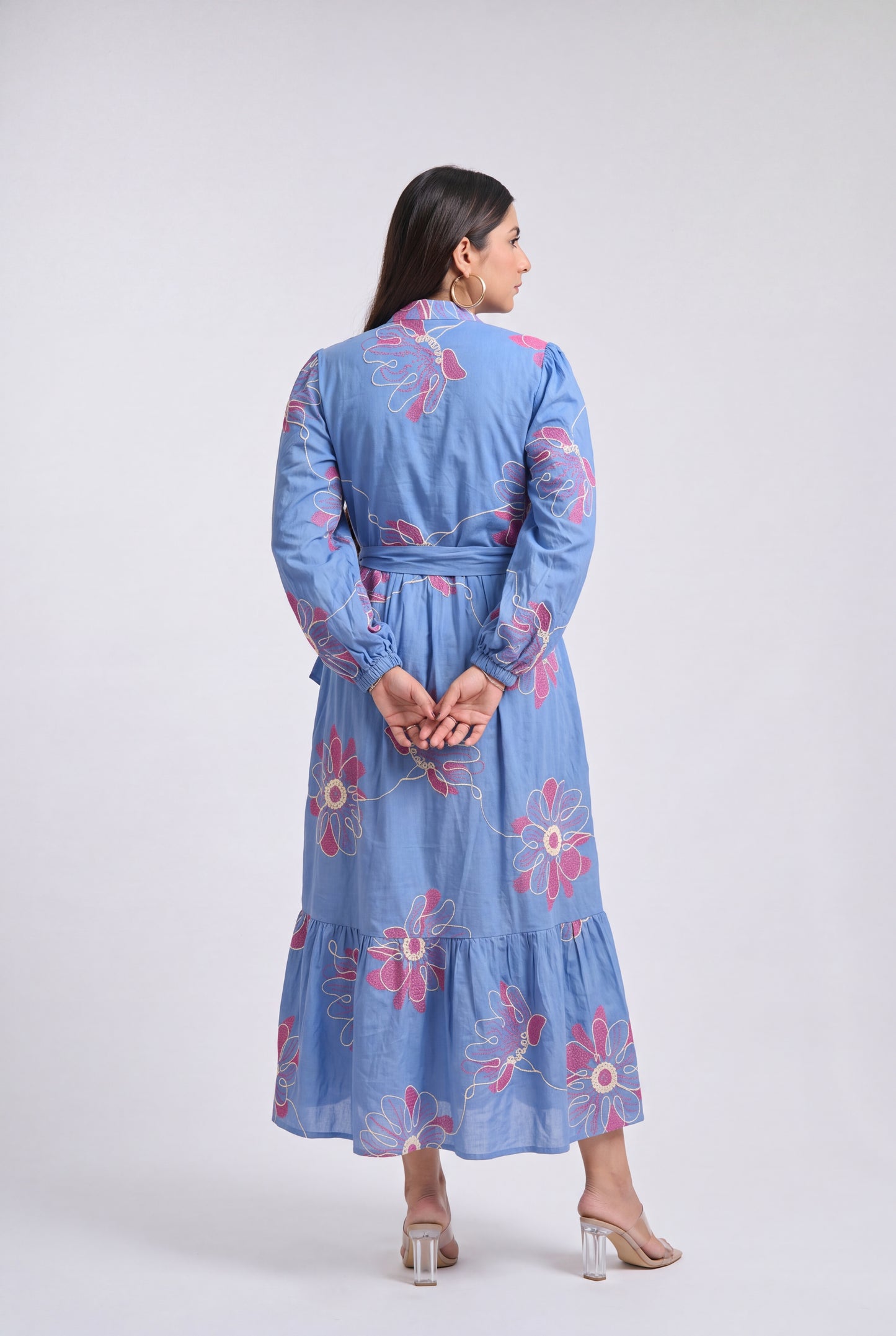 A woman viewed from the back wearing a light blue long-sleeve midi dress with pink floral embroidery, standing against a plain light gray studio background.