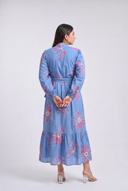 A woman viewed from the back wearing a light blue long-sleeve midi dress with pink floral embroidery, standing against a plain light gray studio background.
