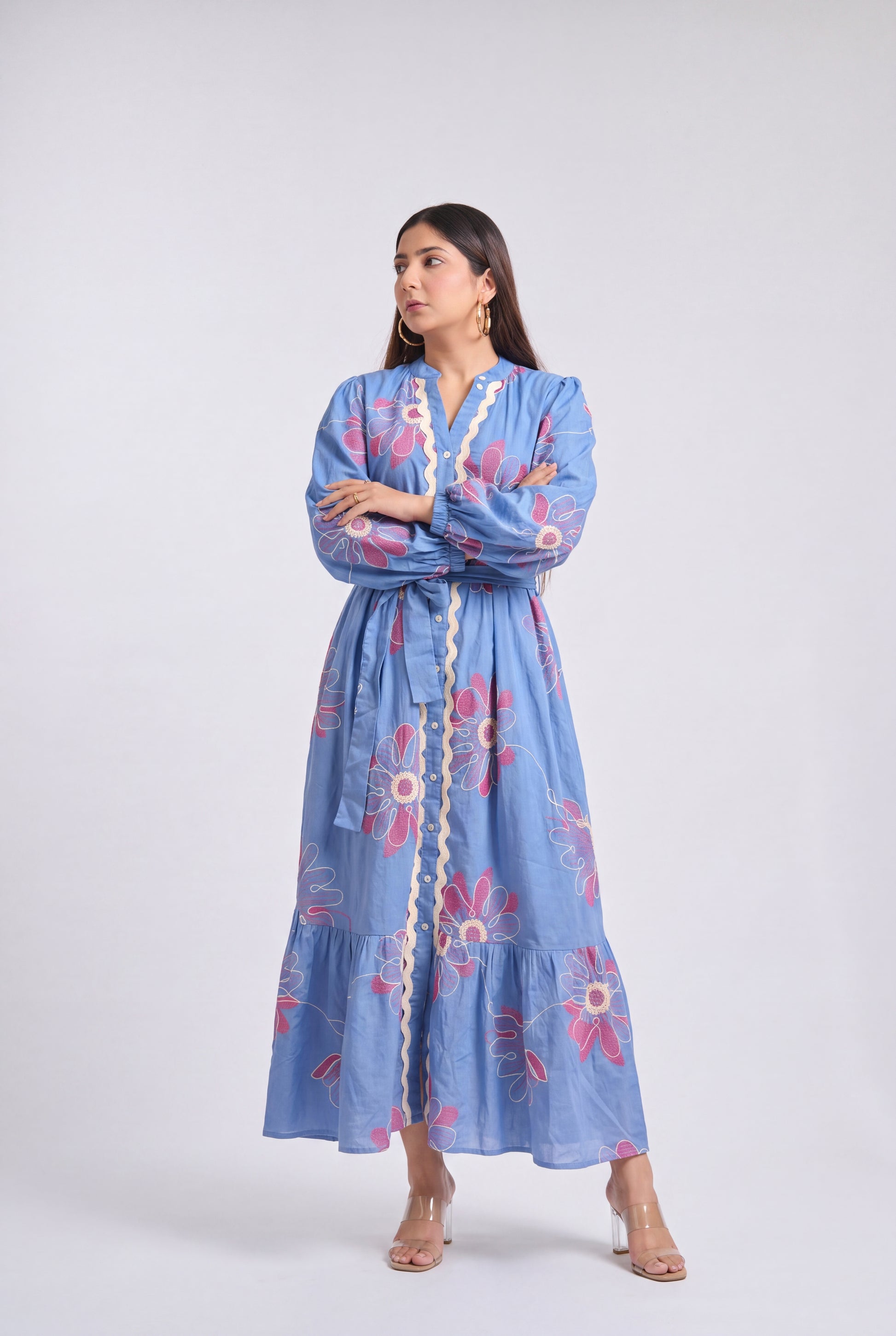 A woman modeling a light blue long-sleeve maxi dress with pink and white floral embroidery and a cream-colored button placket, standing against a light gray studio background.