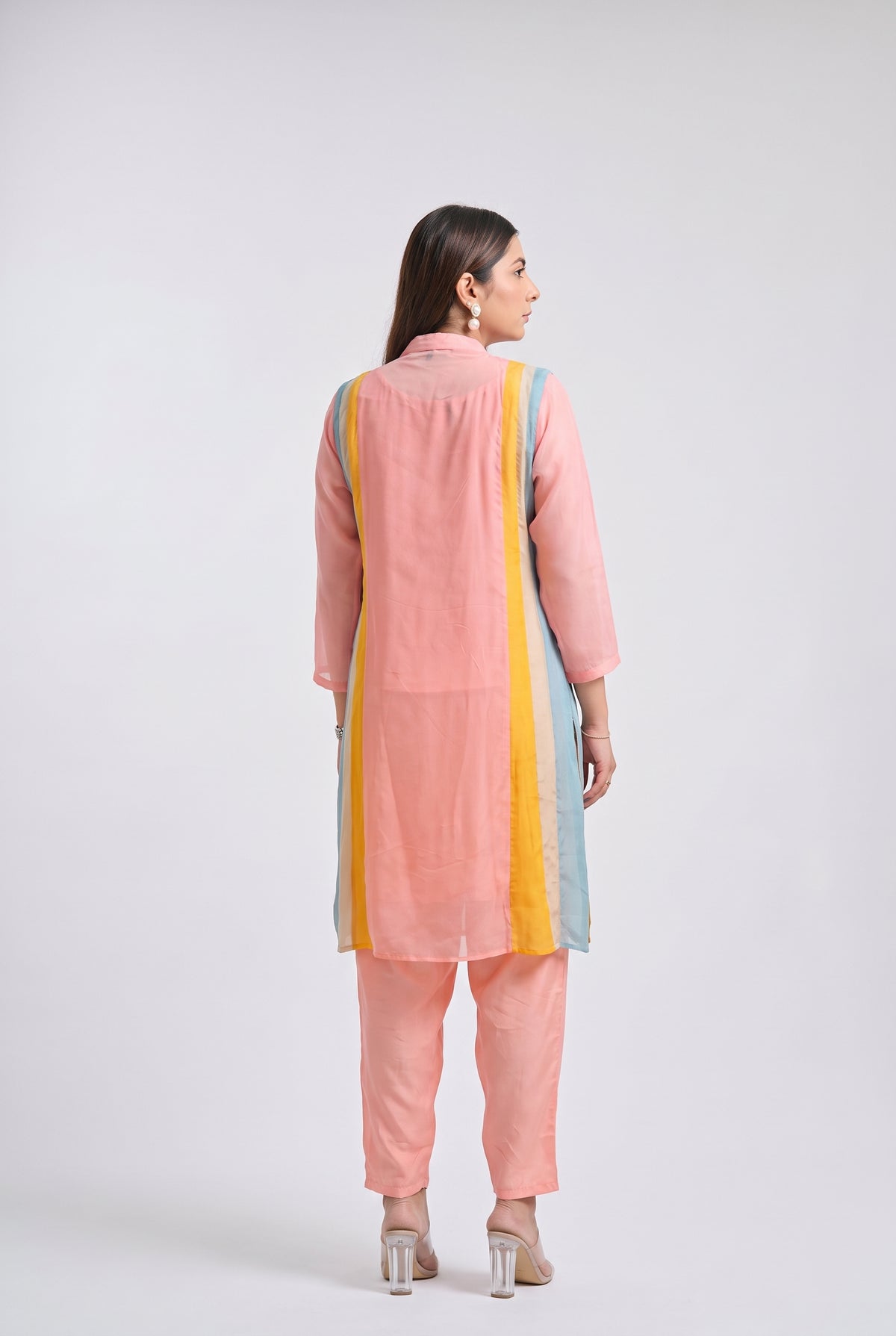 A woman modeling a light pink kurta set with pastel blue and yellow side panels, a colorful embroidered neckline, matching pink pants, and clear high-heeled sandals, posed against a light gray studio background.