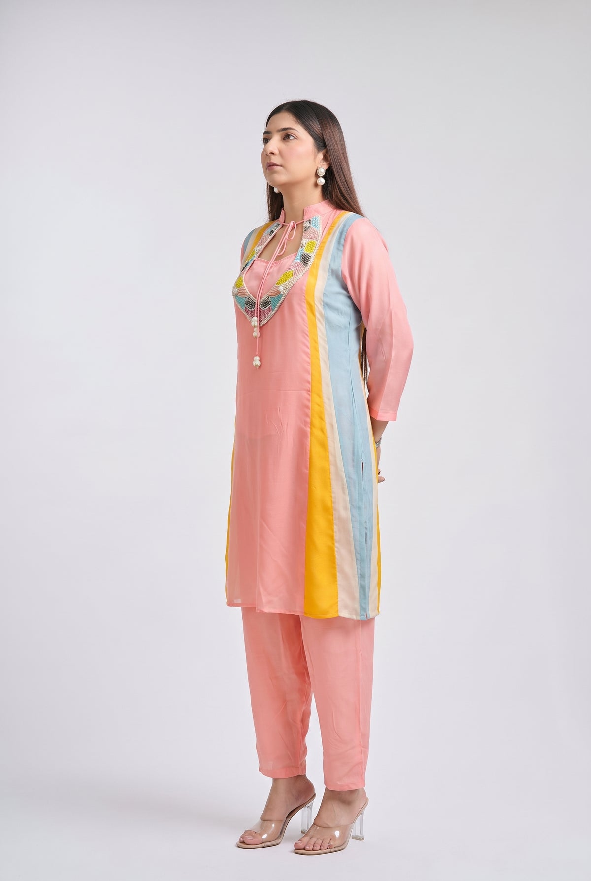 A woman modeling a light pink kurta set with pastel blue and yellow side panels, a colorful embroidered neckline, matching pink pants, and clear high-heeled sandals, posed against a light gray studio background.