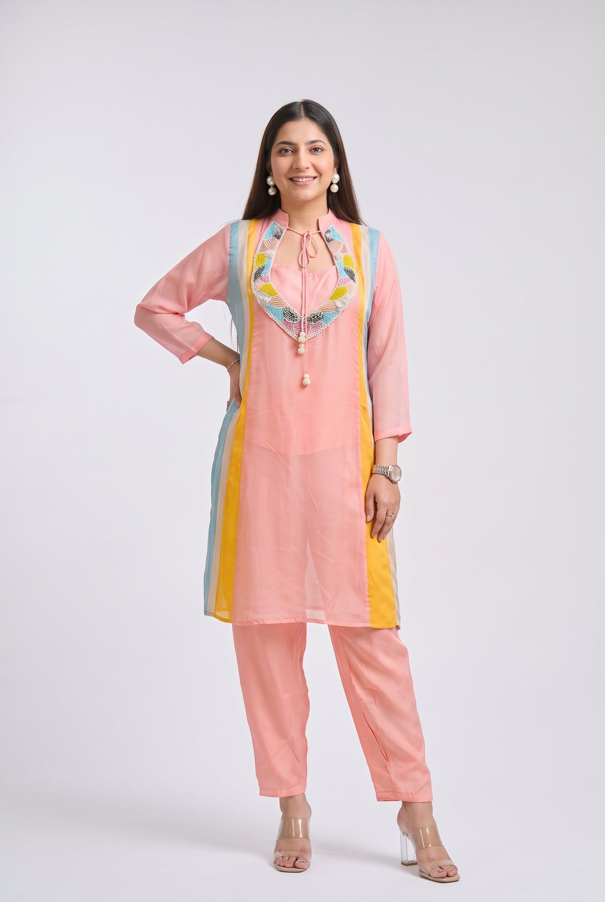 A woman modeling a light pink traditional kurta set with matching pants, featuring yellow and light blue side panels and a colorful embroidered neckline, set against a plain light gray background.