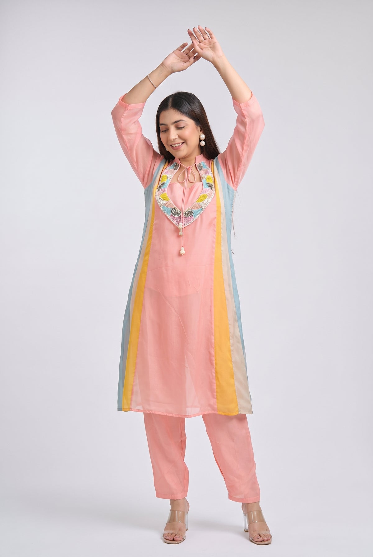 A woman modeling a light pink kurta set with pastel blue and yellow side panels, a colorful embroidered neckline, matching pink pants, and clear high-heeled sandals, posed against a light gray studio background.