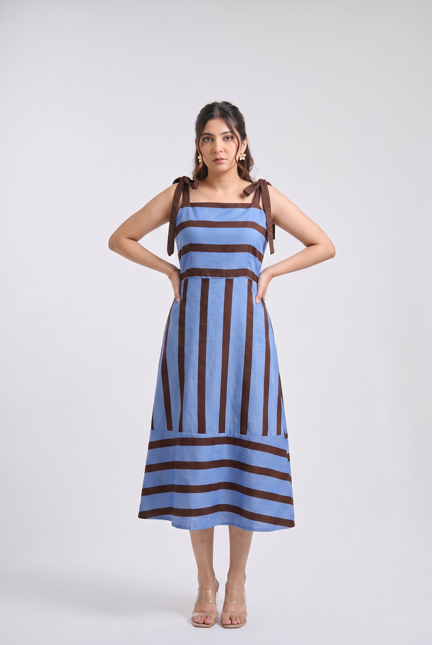 A woman modeling a sleeveless midi dress with vertical blue and dark brown stripes, standing against a light gray studio background.