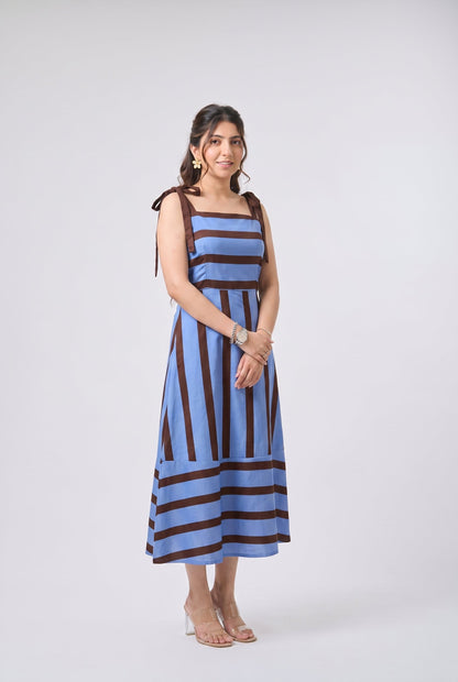 A woman modeling a sleeveless midi dress with vertical blue and dark brown stripes, standing against a light gray studio background.