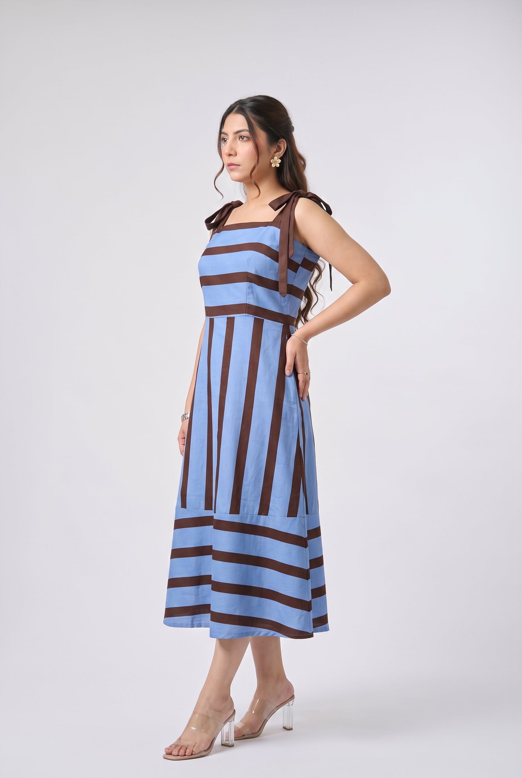 A woman modeling a sleeveless midi dress with vertical light blue and dark brown stripes, featuring tie-shoulder straps and a fitted waist, posed against a light gray studio background.