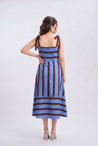 A woman viewed from the back wearing a sleeveless midi dress with vertical dark brown and light blue stripes, tied shoulder straps, and clear high-heeled sandals, against a plain light gray studio background.