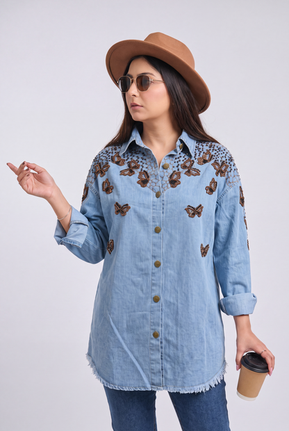 A woman wearing a light blue denim button-up shirt with brown butterfly embroidery on the chest and shoulders, paired with a brown hat, sunglasses, and jeans, holding a coffee cup against a light gray background.