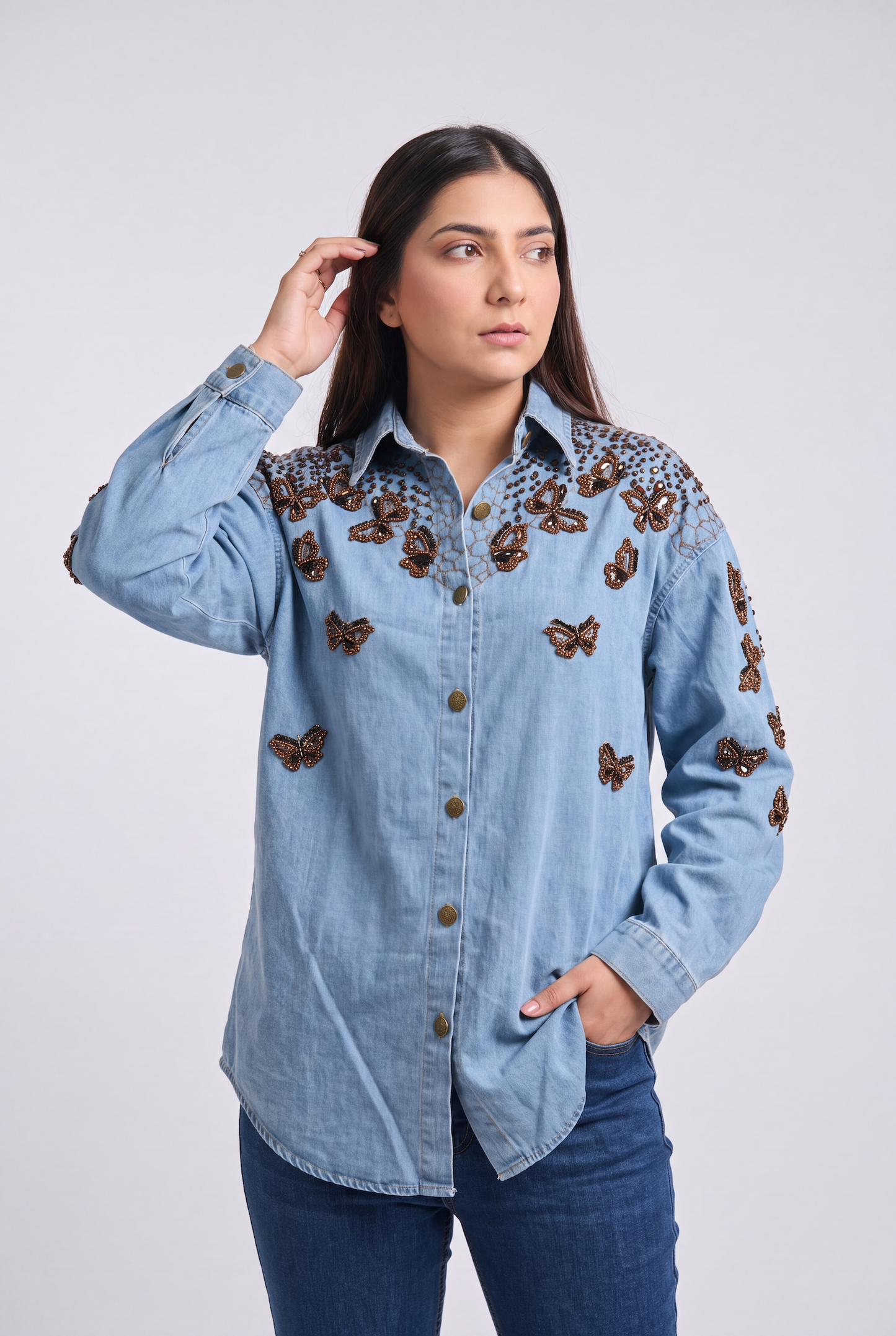 A woman modeling a light blue denim button-up shirt with brown butterfly embroidery on the shoulders and chest, paired with dark blue jeans against a light gray studio background.