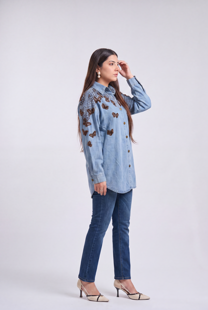 A woman modeling a light blue denim shirt with brown butterfly embroidery, paired with dark blue jeans and beige heels, standing against a light gray studio background.