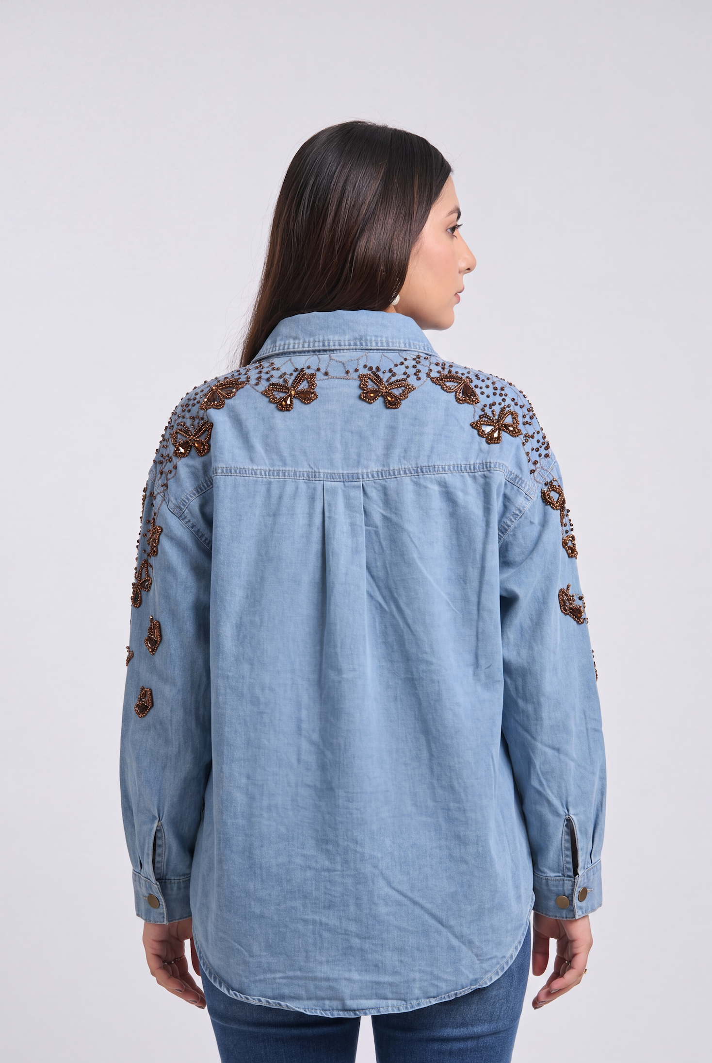A woman viewed from the back wearing a light blue denim shirt jacket with brown embroidered floral details across the shoulders and upper sleeves, paired with dark blue jeans against a light gray studio background.
