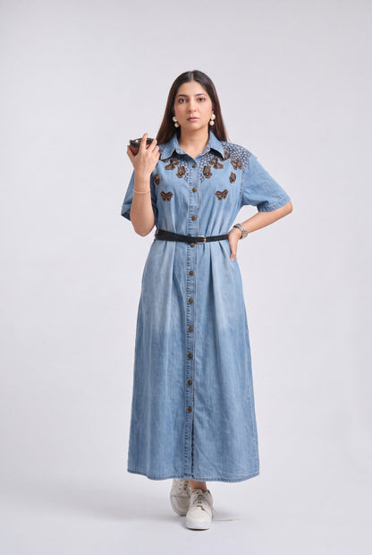 A woman modeling a light blue denim midi dress with butterfly embroidery on the chest, a black belt, and white sneakers, standing against a light gray studio background.