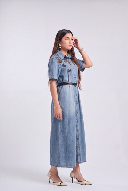 A woman modeling a light blue denim midi dress with butterfly embroidery on the chest, a black belt at the waist, and button-front detailing, photographed against a plain light gray studio background.