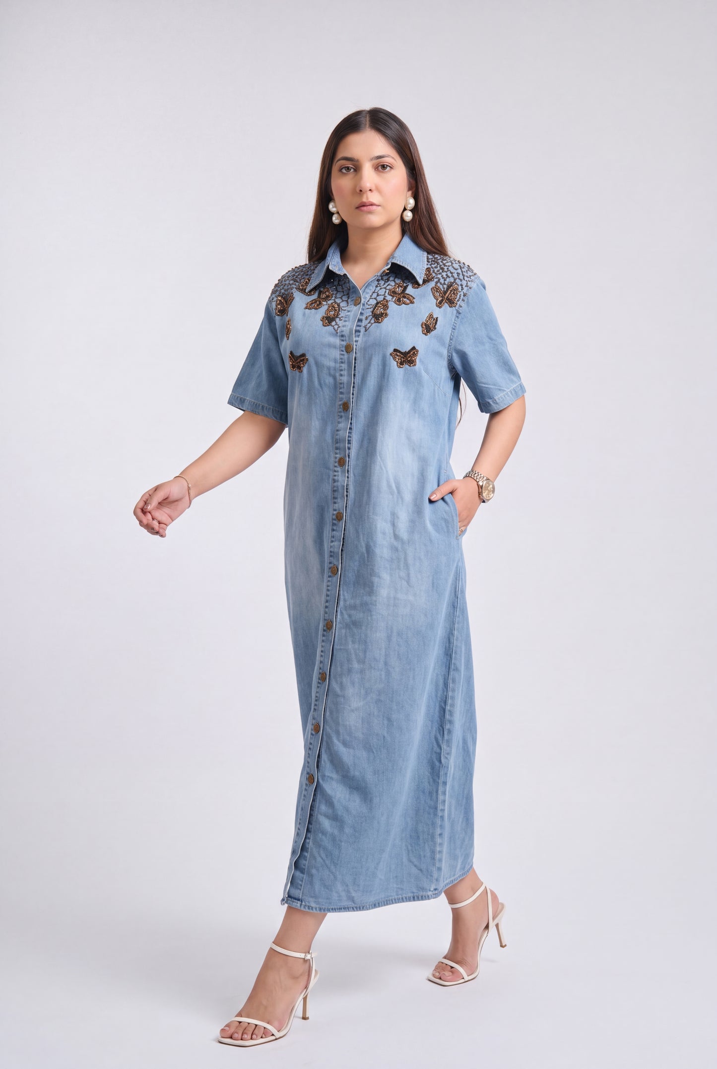 A woman modeling a light blue denim maxi dress with brown embroidered butterfly details on the chest, standing against a light gray studio background.