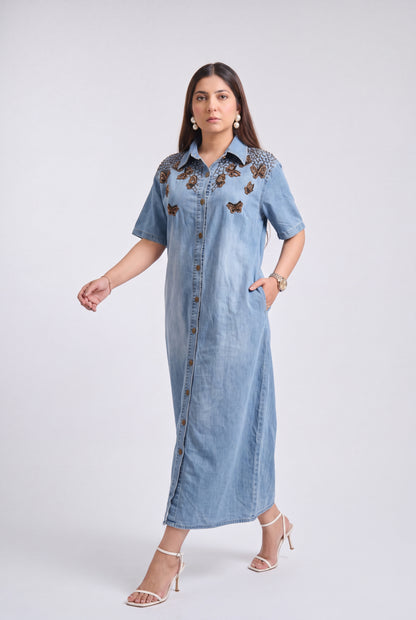 A woman modeling a light blue denim maxi dress with brown embroidered butterfly details on the chest, standing against a light gray studio background.