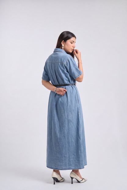 A woman modeling a light blue denim maxi dress with short sleeves and a thin black belt, shown from a side-back angle against a plain light gray studio background.