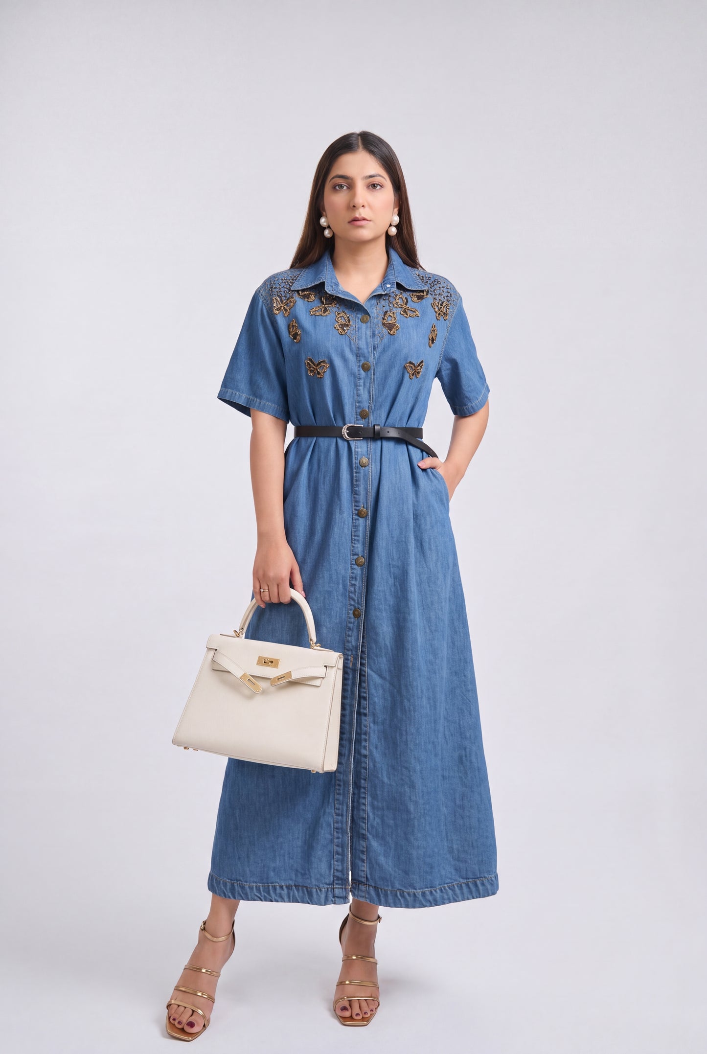 A woman modeling a blue denim midi dress with gold and brown butterfly embroidery on the chest, styled with a black belt and holding a cream-colored handbag against a light gray studio background.