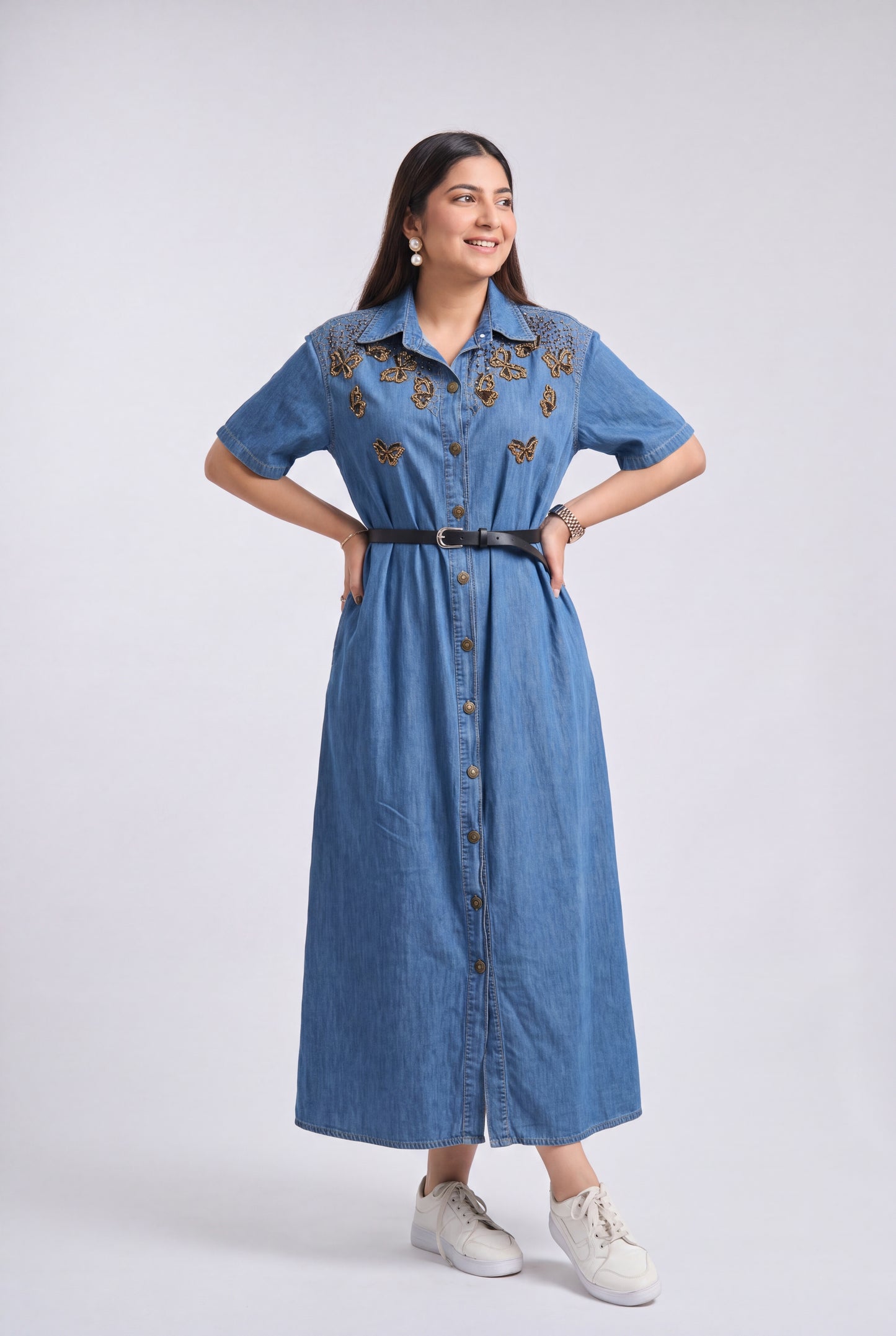 A woman modeling a blue denim midi dress with butterfly embroidery on the chest, a black belt, and white sneakers, photographed against a light gray studio background.