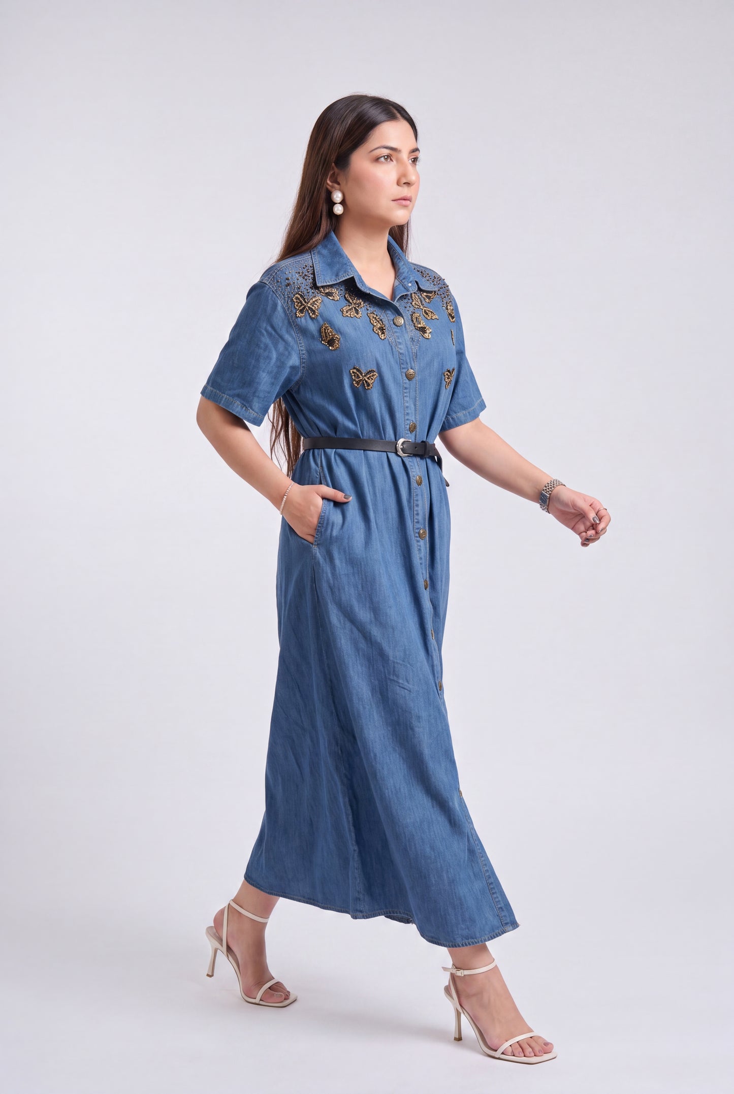 A woman modeling a blue denim midi dress with gold butterfly embroidery on the chest, a black belt, and beige high-heeled sandals against a light gray studio background.