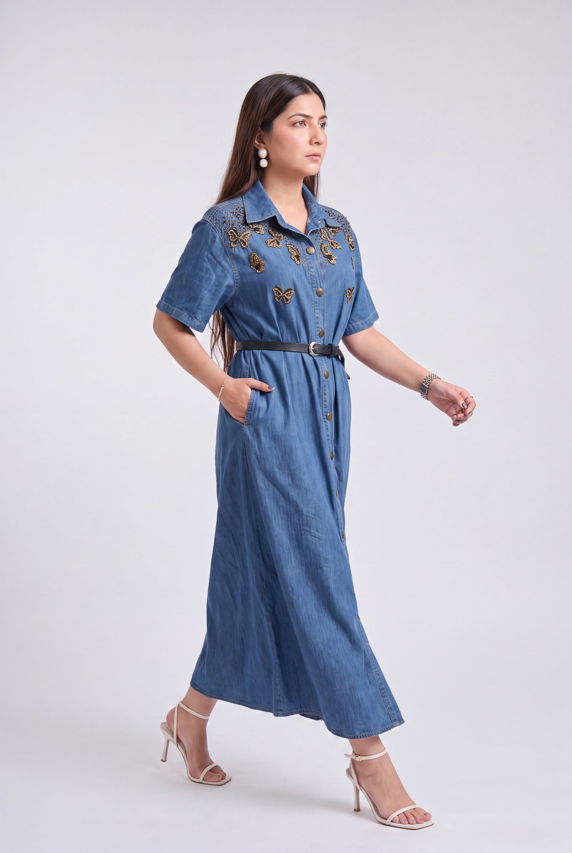 A woman modeling a blue denim midi dress with gold butterfly embroidery on the chest, a black belt, and beige high-heeled sandals against a light gray studio background.