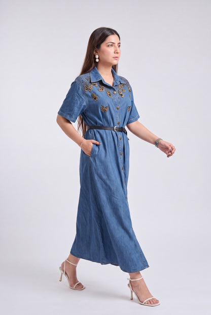A woman modeling a blue denim midi dress with gold butterfly embroidery on the chest, a black belt, and beige high-heeled sandals against a light gray studio background.
