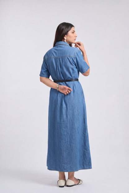 A woman wearing a blue denim midi dress with short sleeves, a high collar, and a thin black belt at the waist, shown from a rear three-quarter view against a light gray studio background.