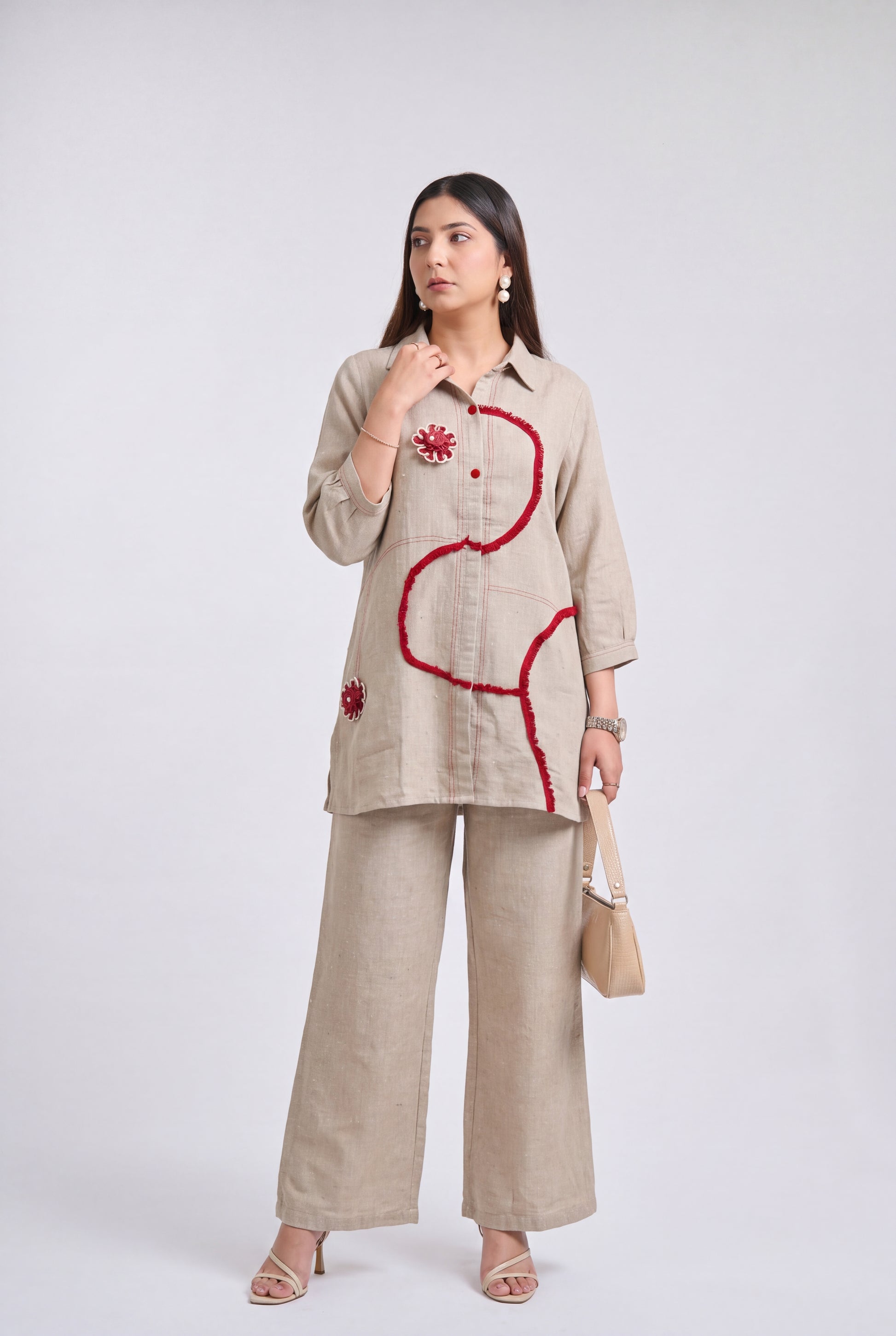 A woman models a beige linen two-piece set consisting of a long-sleeve button-up shirt with red sequined embroidery and matching wide-leg trousers, holding a beige handbag against a light gray studio background.