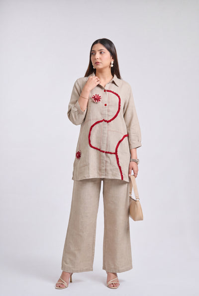 A woman models a beige linen two-piece set consisting of a long-sleeve button-up shirt with red sequined embroidery and matching wide-leg trousers, holding a beige handbag against a light gray studio background.