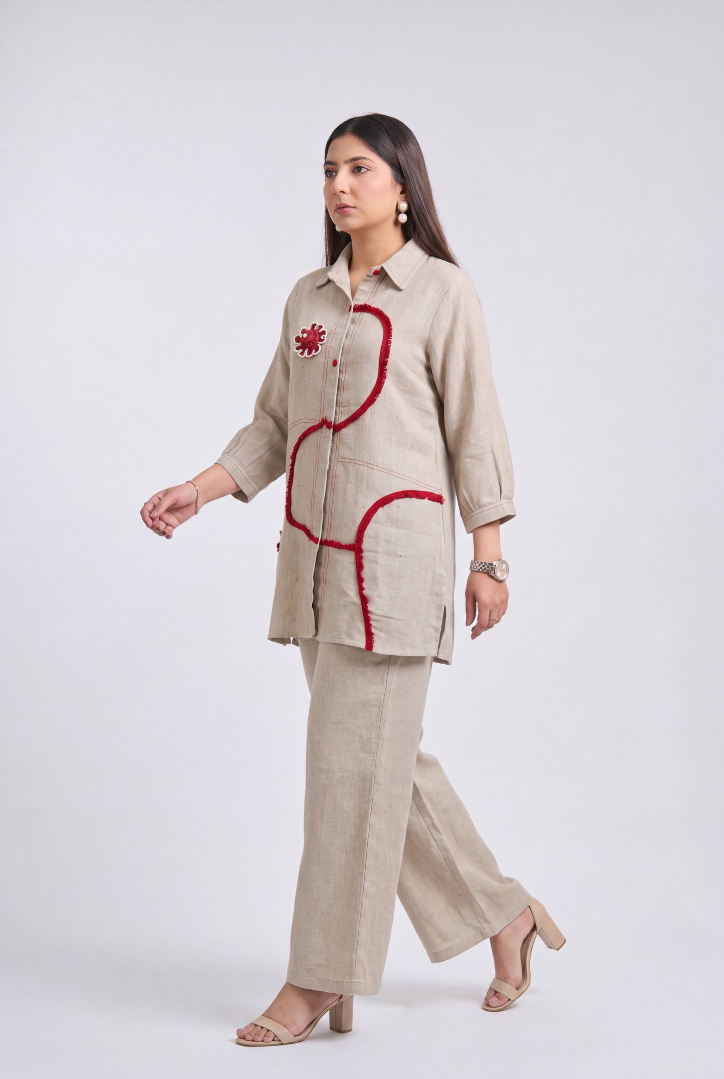 A woman modeling a beige linen two-piece set consisting of a long-sleeve button-up shirt and matching wide-leg trousers, featuring red embroidered accents on the shirt.