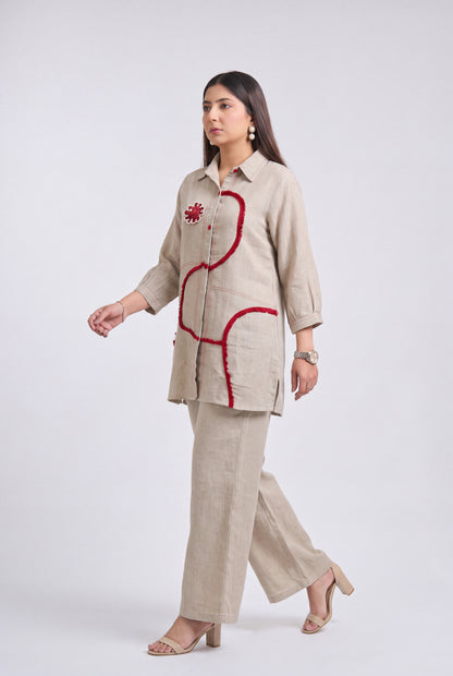 A woman modeling a beige linen two-piece set consisting of a long-sleeve button-up shirt and matching wide-leg trousers, featuring red embroidered accents on the shirt.