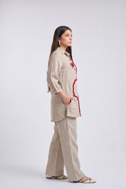 A woman stands in profile wearing a light beige linen two-piece outfit with red embroidered details on the jacket and pants, paired with beige flat sandals, against a plain light gray studio background.