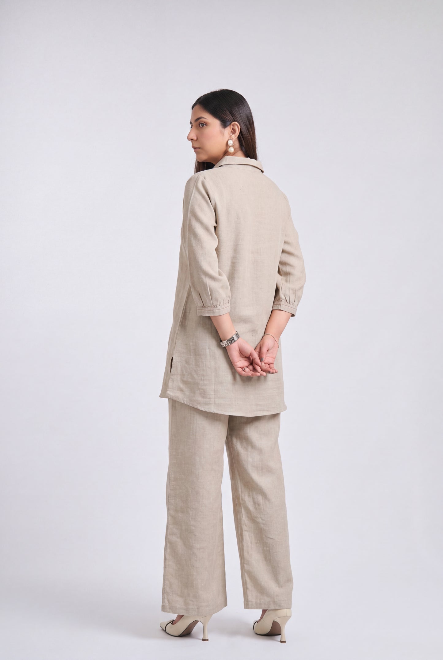 A woman modeling a light beige linen two-piece set consisting of a long-sleeve tunic and wide-leg trousers, shown from a rear three-quarter view against a light gray studio background.