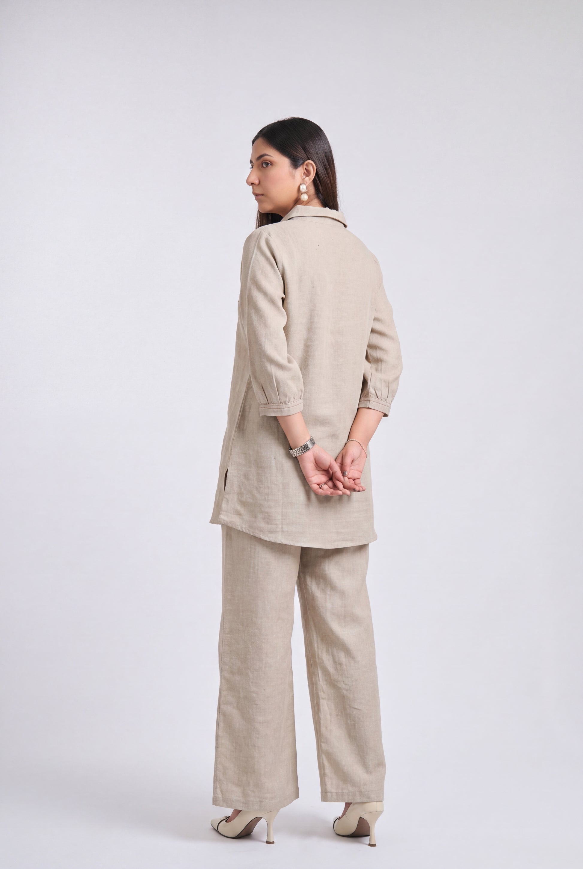 A woman modeling a light beige linen two-piece set consisting of a long-sleeve tunic and wide-leg trousers, shown from a rear three-quarter view against a light gray studio background.