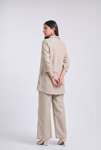 A woman modeling a light beige linen two-piece set consisting of a long-sleeve tunic and wide-leg trousers, shown from a rear three-quarter view against a light gray studio background.