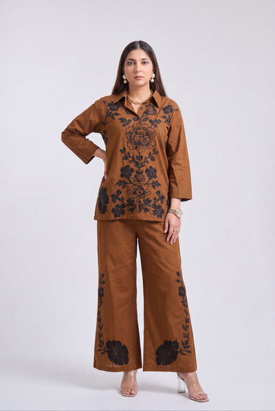 A woman modeling a matching brown two-piece outfit with black floral embroidery, consisting of a long-sleeve button-up top and wide-leg pants, against a light gray studio background.