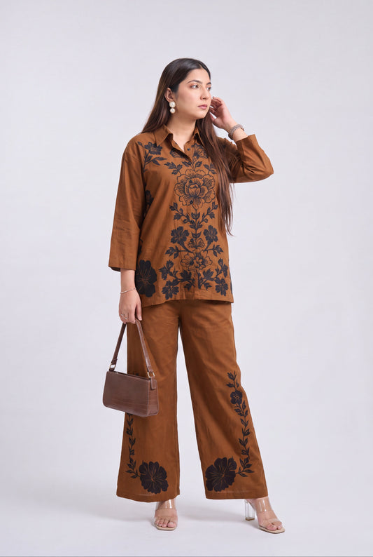 A woman modeling a two-piece brown pajama set with black floral embroidery, featuring a long-sleeve button-up top and matching wide-leg pants, styled with a brown handbag against a light gray background.