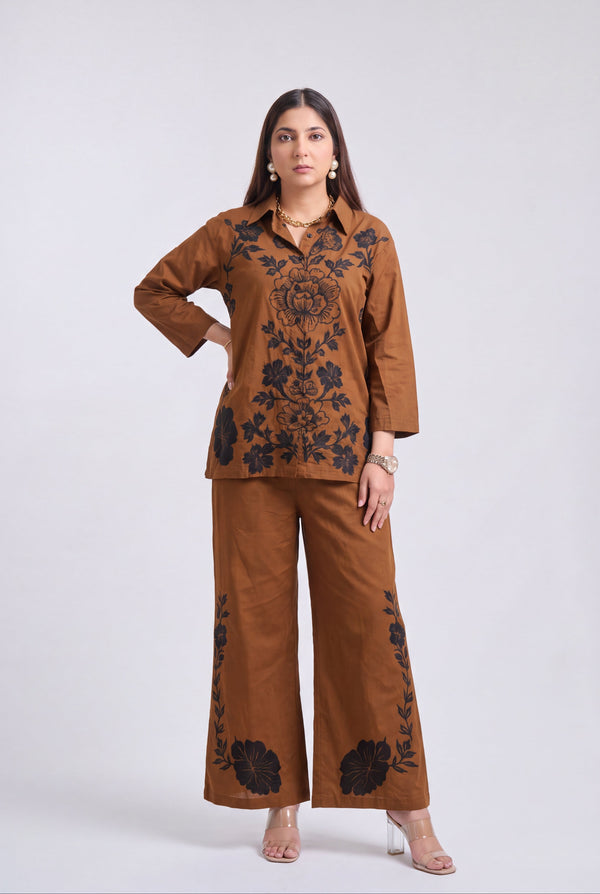 A woman modeling a matching brown two-piece outfit with black floral embroidery, consisting of a long-sleeve button-up top and wide-leg pants, against a light gray studio background.