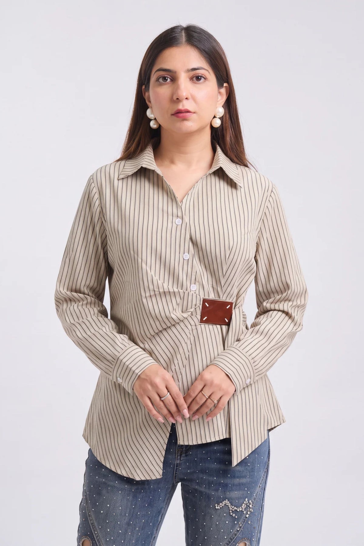 A woman modeling a beige and brown vertically striped long-sleeve button-up shirt with a small brown leather detail at the waist, paired with blue jeans, against a light gray background.