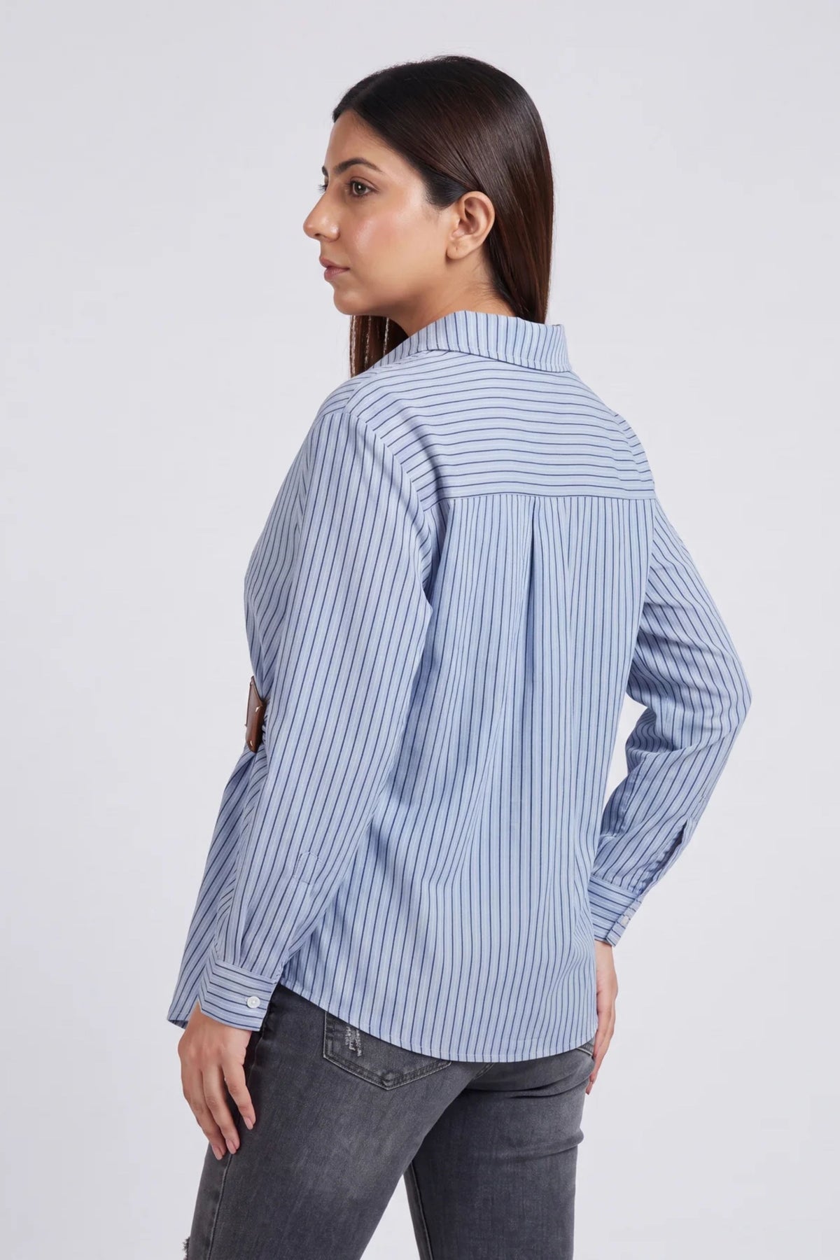 A woman shown from the back wearing a light blue and white vertically striped button-up shirt with dark gray jeans, photographed against a plain light gray studio background.
