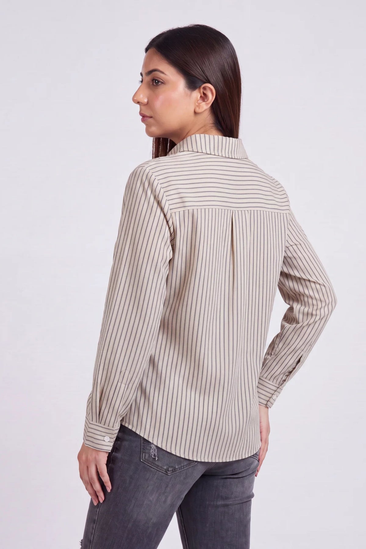 A woman shown from the back wearing a beige and white vertically striped long-sleeve blouse with dark gray jeans, photographed against a light gray studio background.