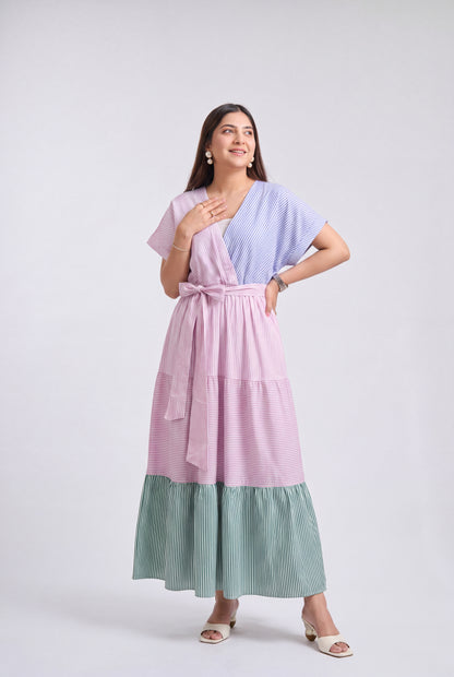 A woman modeling a three-tiered wrap maxi dress with a light purple upper section, a pink tiered skirt, and a sage green striped bottom, set against a plain light gray studio background.
