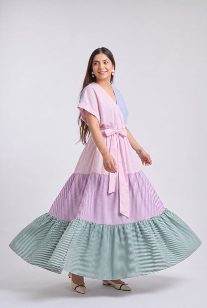 A woman modeling a tiered maxi dress with a color-blocked design featuring light pink, lavender purple, and mint green pinstripes, set against a plain light gray studio background.