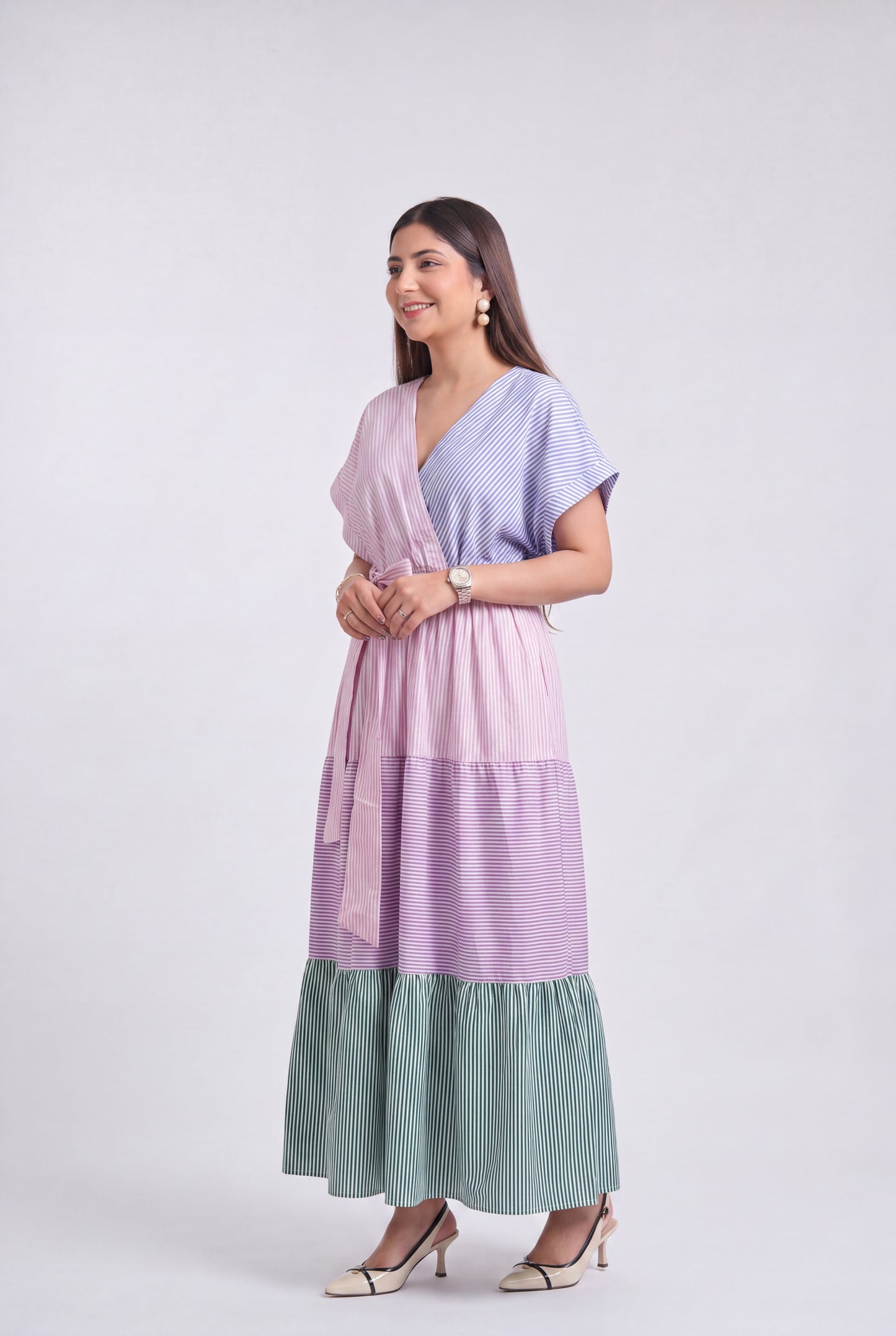 A woman modeling a long, tiered wrap dress with a lavender and light blue striped pattern and a sage green pleated ruffled hem, set against a plain light gray studio background.