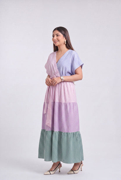 A woman modeling a long, tiered wrap dress with a lavender and light blue striped pattern and a sage green pleated ruffled hem, set against a plain light gray studio background.