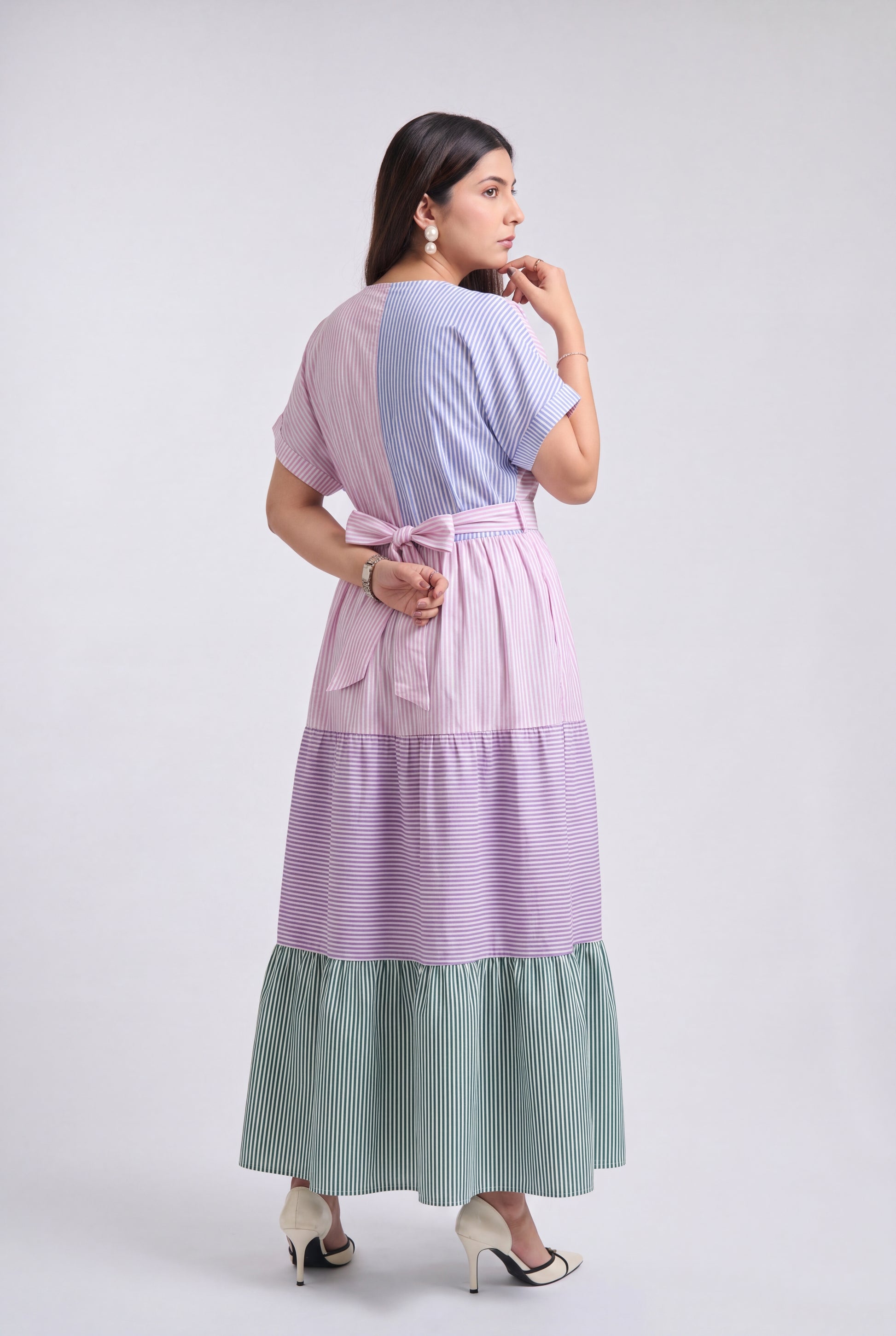 A woman modeling a long tiered maxi dress with horizontal color-blocked panels in lavender, light purple, and sage green, shown from the back against a light gray studio background.