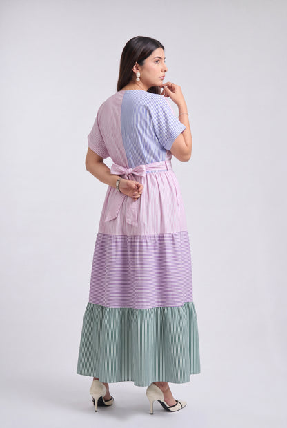 A woman modeling a long tiered maxi dress with horizontal color-blocked panels in lavender, light purple, and sage green, shown from the back against a light gray studio background.