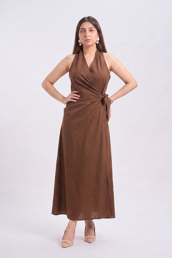 A woman modeling a sleeveless brown halter maxi dress with a wrap-style bodice and side tie, paired with beige heels against a plain light gray studio background.