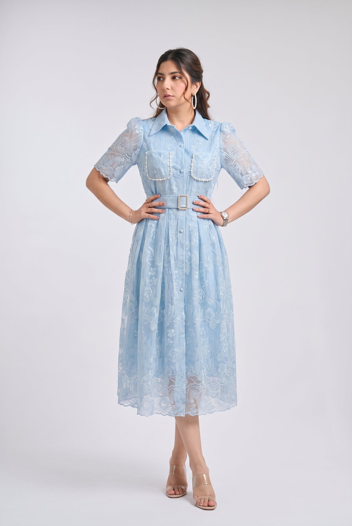A woman modeling a light blue midi dress with sheer lace short sleeves, a button-up front, decorative chest pockets with pearl trim, and a matching lace belt with a gold buckle, set against a plain light gray studio background.