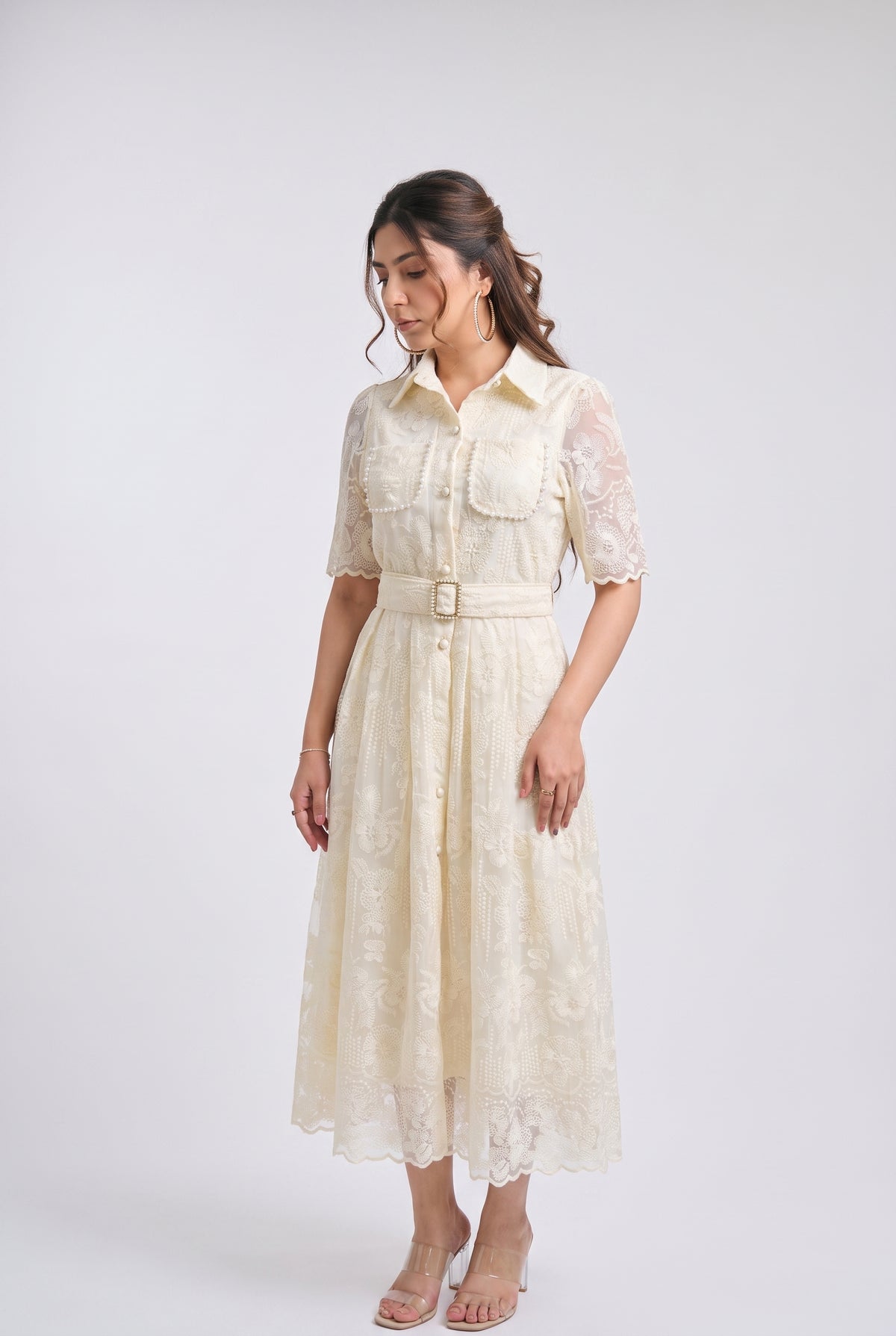 A woman modeling a cream-colored midi dress with sheer lace sleeves, a button-up front, and a matching fabric belt with a gold-tone buckle, set against a plain light gray studio background.